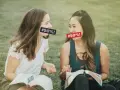 Two women with censored mouths discuss if cursing is a sin while reading books on the grass.
