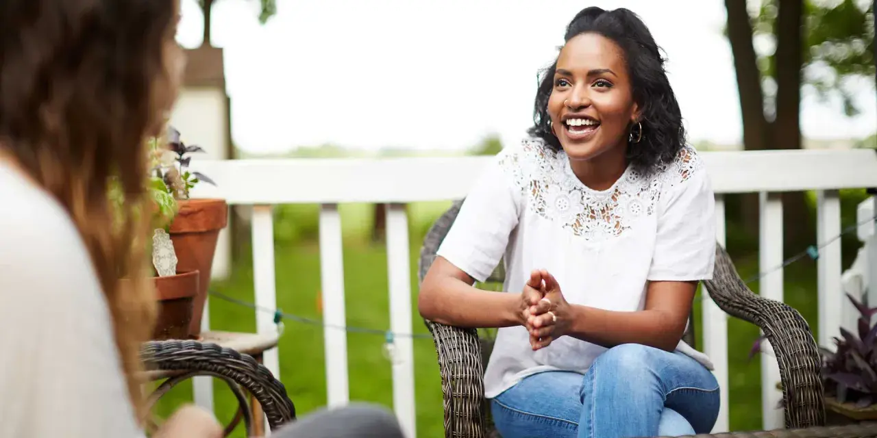 Two women are sitting on a porch, one animatedly sharing the gospel with the other.