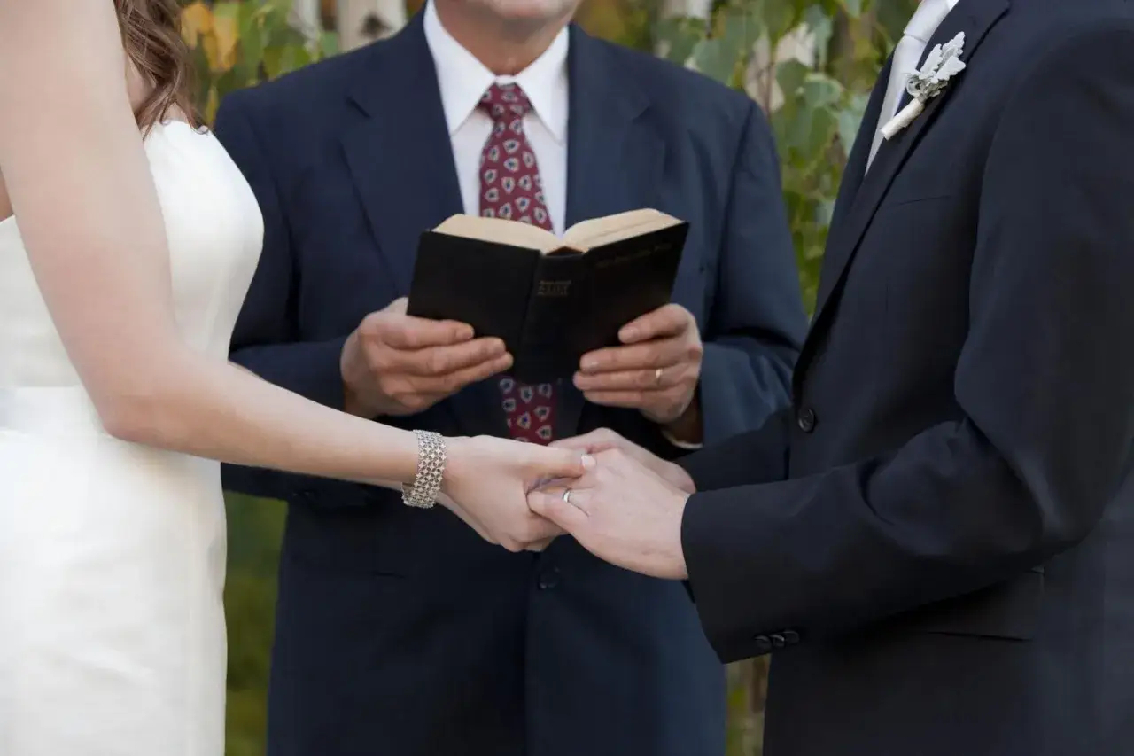 A wedding ceremony, with the officiant holding a book and the couple's hands clasped in a symbolic laying on of hands.