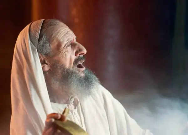 An elderly man, possibly Zechariah in the Bible, with a grey beard and wearing a white robe and head covering, cries out with his mouth open.